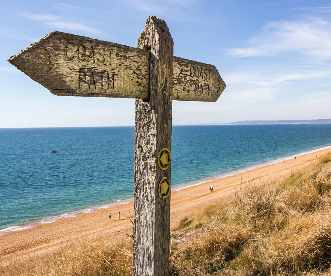 Wooden signpost on a coastal path, with arrows pointing toward Blackbird Cottage and sandy beach under a clear blue sky.