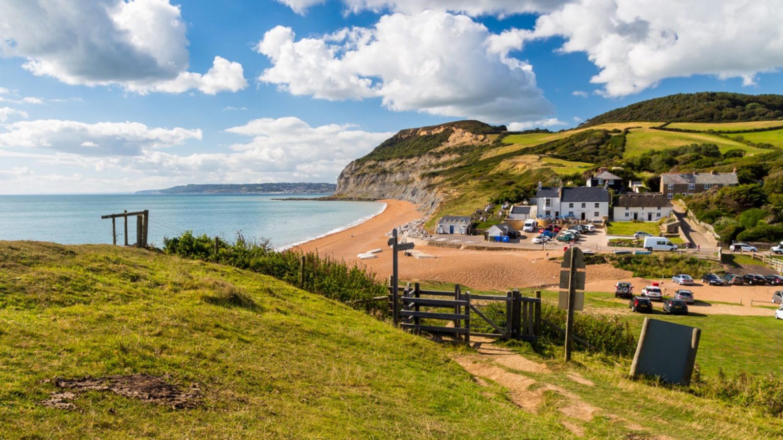 A coastal scene with sandy beach, ocean, grassy cliffs, and Blackbird Cottage in a small village under partly cloudy sky.