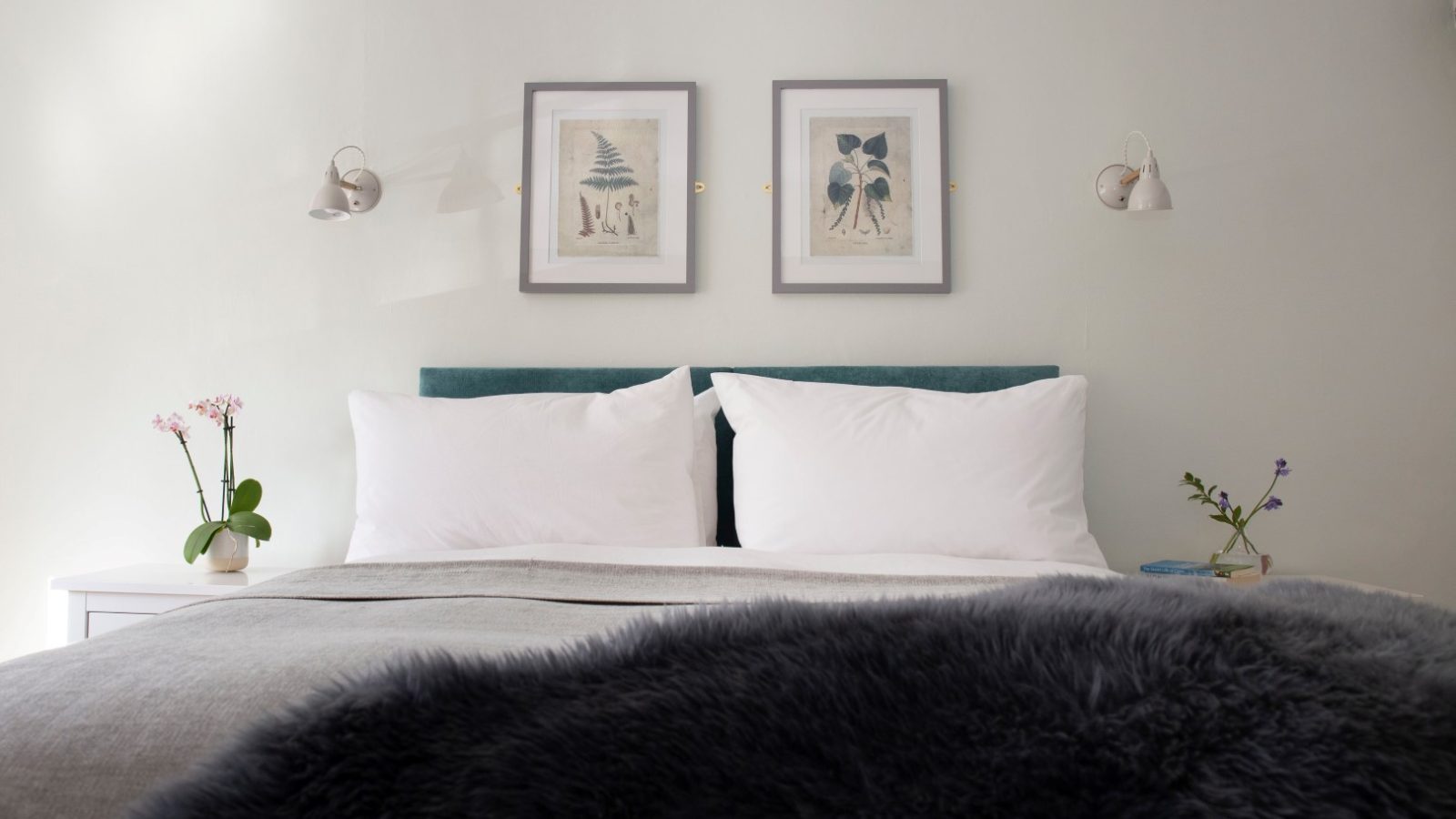 A neatly made bed with white pillows and a gray blanket. Two framed botanical prints add charm to Blackbird Cottage.