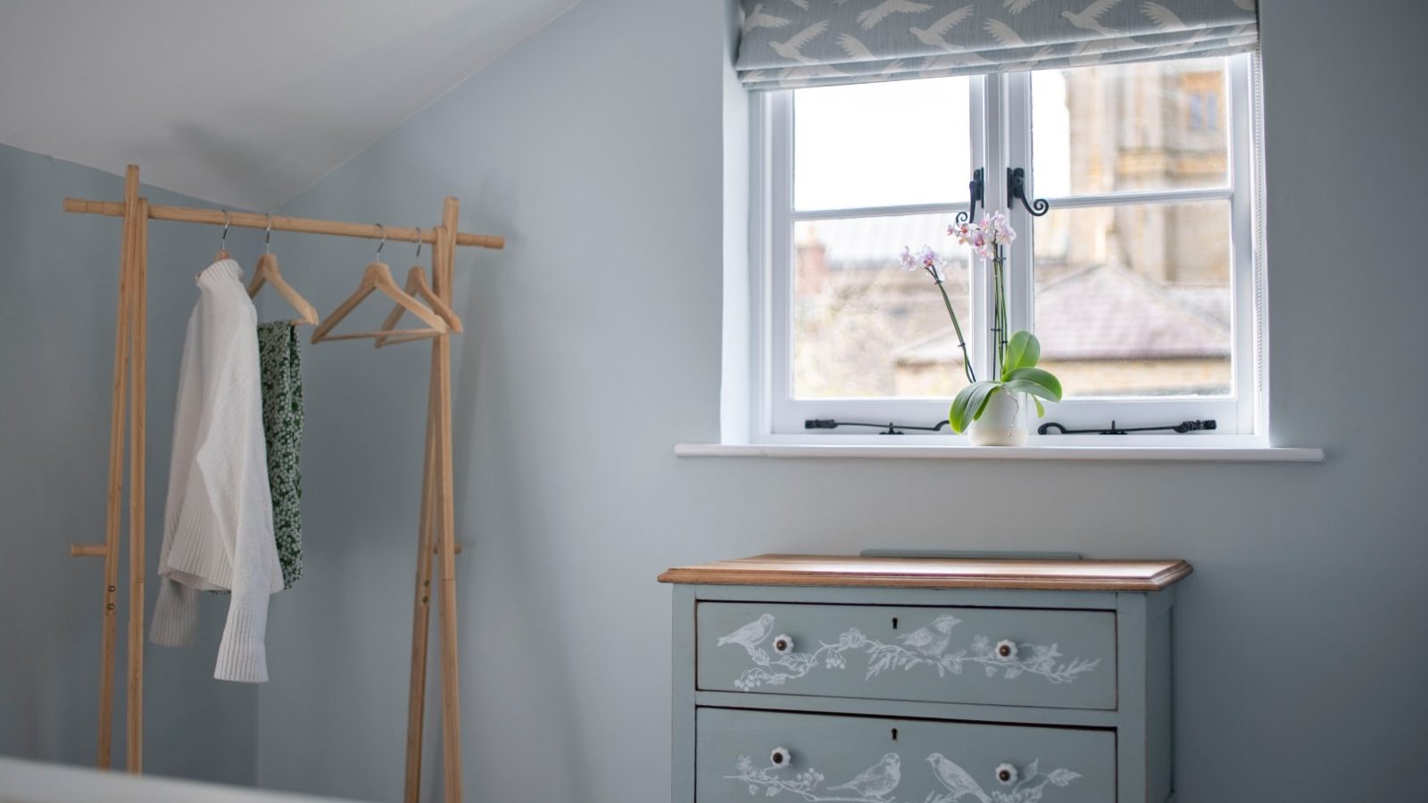 Blackbird Cottage: Light blue bedroom with wooden clothes rack, two hangers, and a chest of drawers under a window with a potted plant.