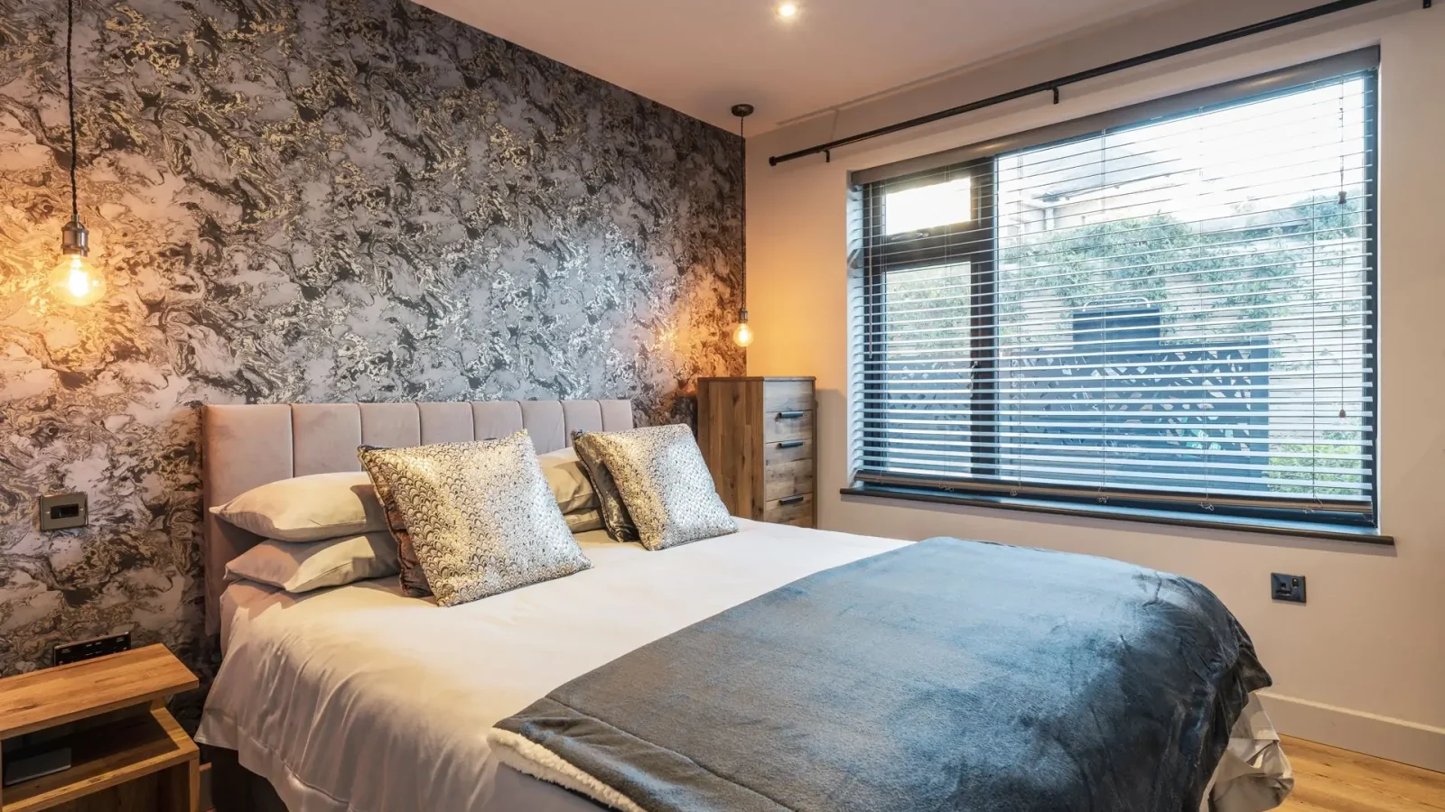 A modern bedroom with a double bed, decorative wallpaper, bedside tables, and blinds offering a serene harbour view.