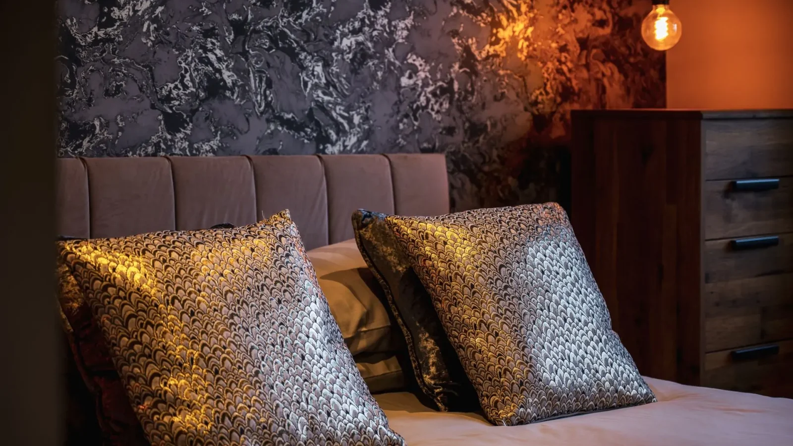 Bedroom with a textured dark wall, two metallic patterned pillows, and a warm glowing lamp harbored on a wooden dresser.