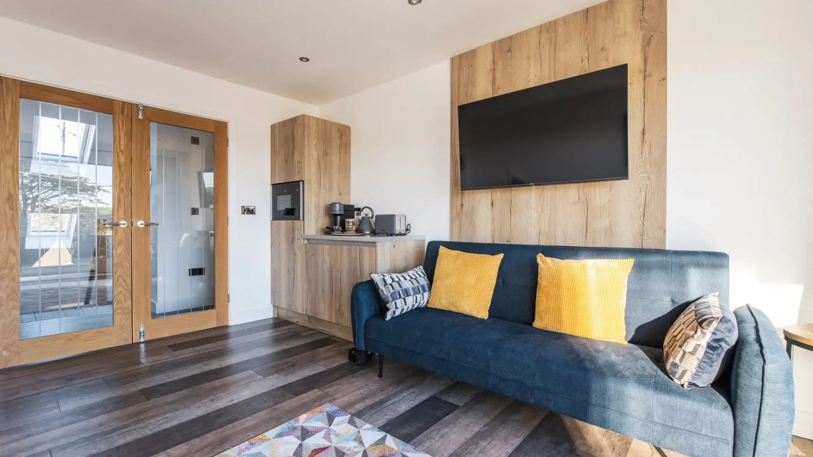 Modern Harbour Reach living room with blue sofa, yellow cushions, wall-mounted TV, kitchenette, and wooden accents throughout.