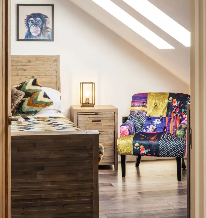 Cozy attic bedroom with wooden bed, colorful armchair, bedside table, and artwork. Skylights brighten Harbour Reach's charm.