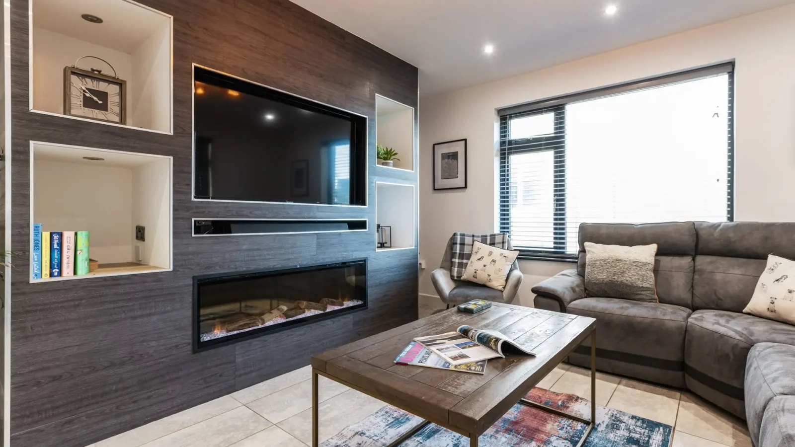 Modern living room with fireplace, gray sectional sofa, and coffee table; a mounted TV reaches over shelves with decor.