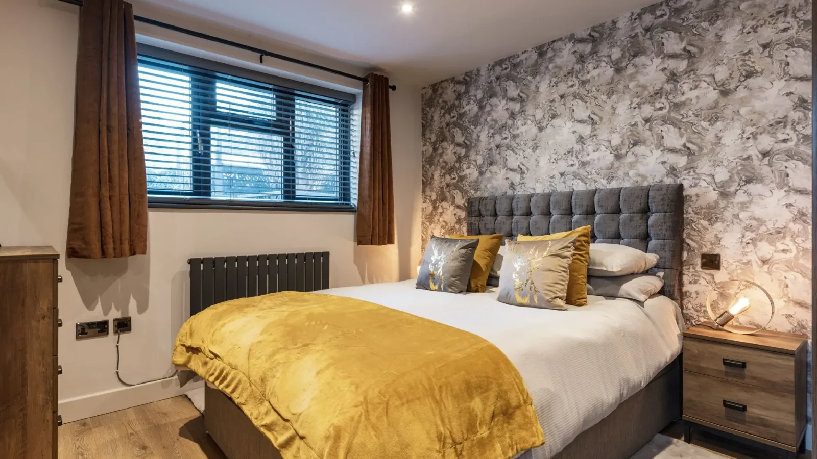 Bedroom with double bed, mustard yellow bedding, wooden nightstands, patterned accent wall, and brown curtains reaching the floor.