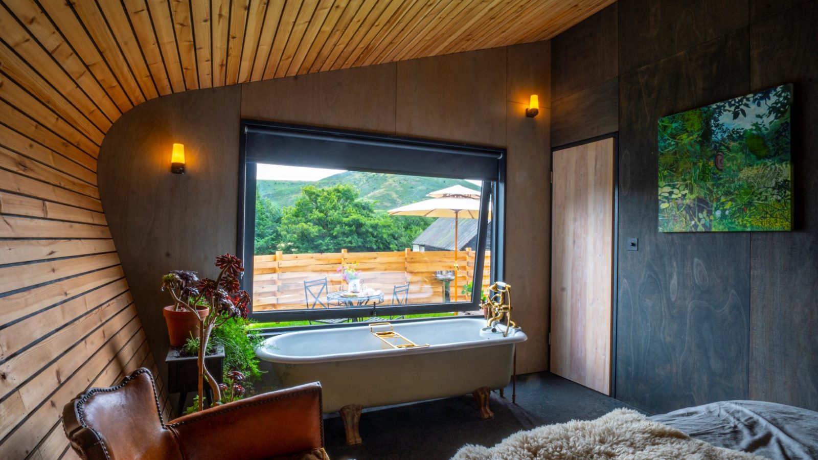 Cozy room with a freestanding bathtub, Hergest Lee leather chair, and a large window overlooking a terrace with outdoor furniture.