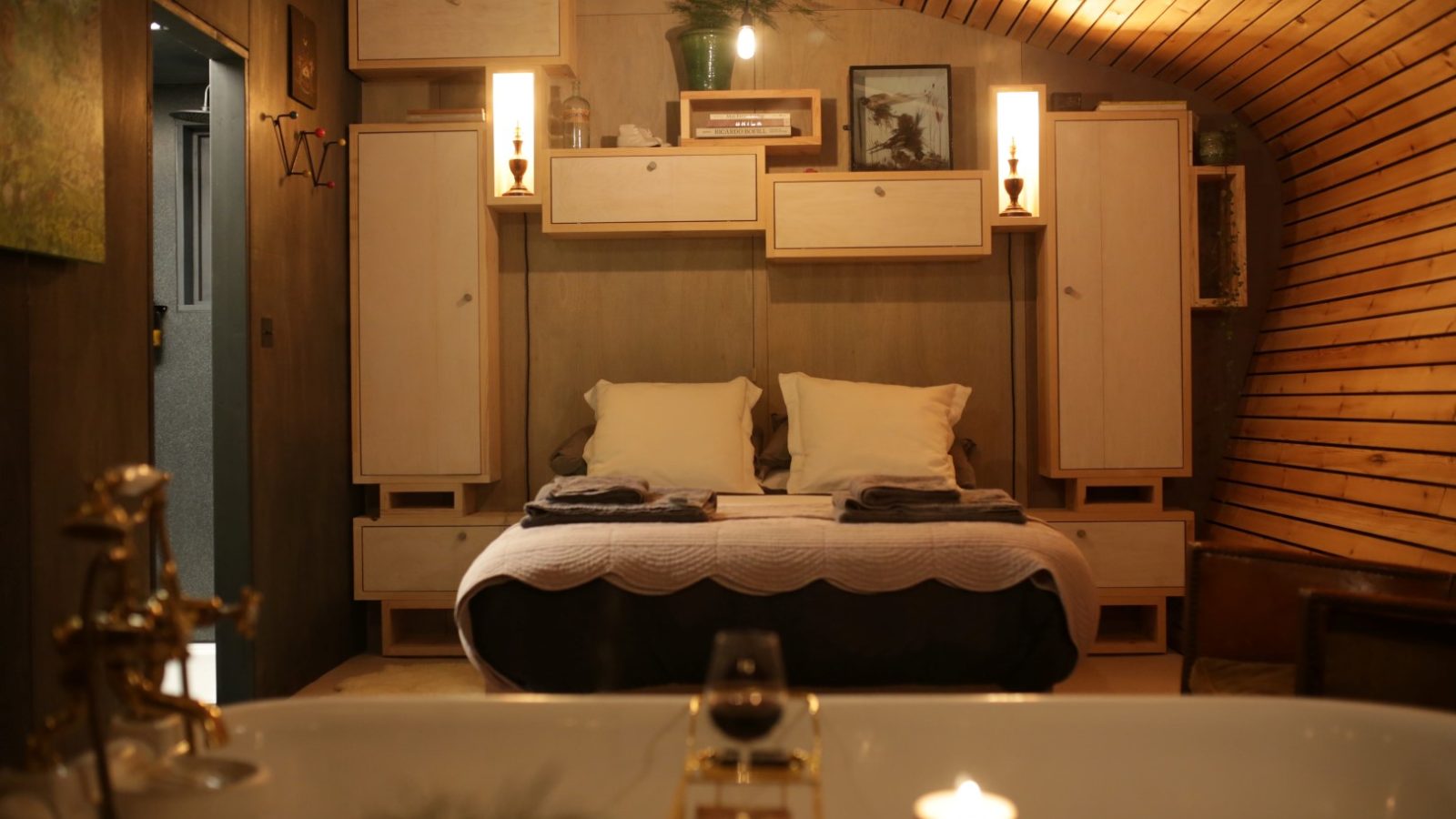 Cozy bedroom with Hergest Lee paneling, a central bed, warm lighting, and a bathtub with a glass of red wine.