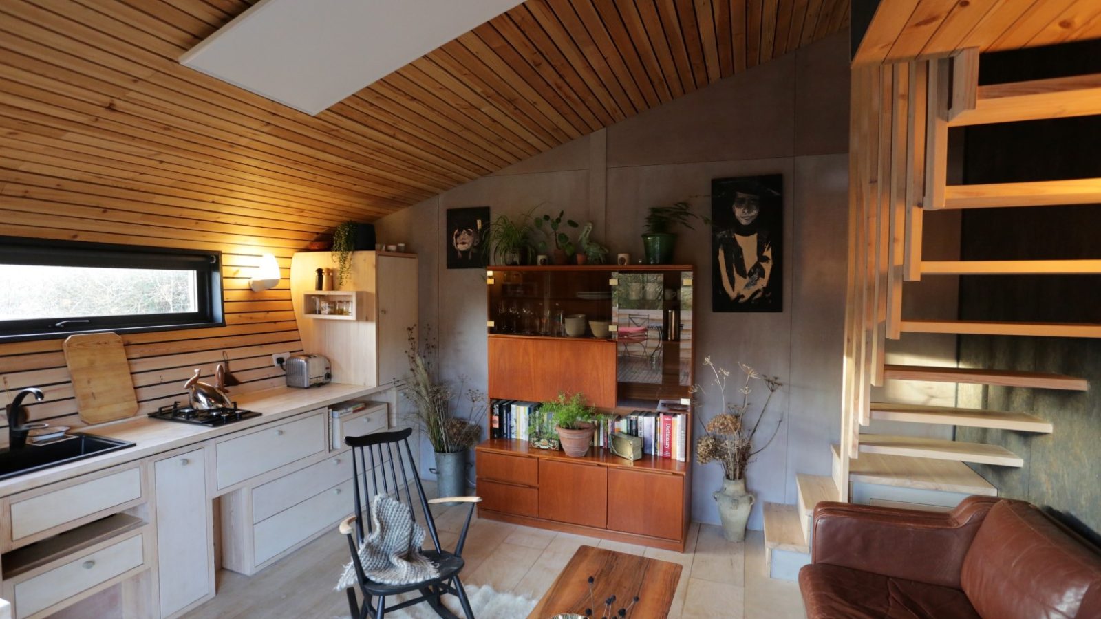 Cozy Hergest Lee living area with wooden ceiling, a kitchenette, rocking chair, bookshelf, and modern staircase.