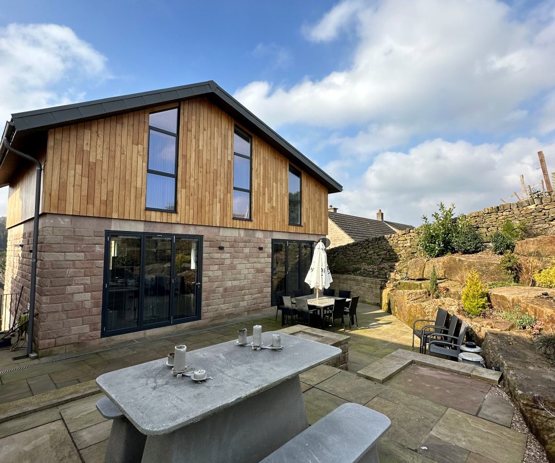 Oakwood House is a modern home with a wooden and stone exterior, featuring a patio complete with a concrete picnic table and benches under a vast blue sky.