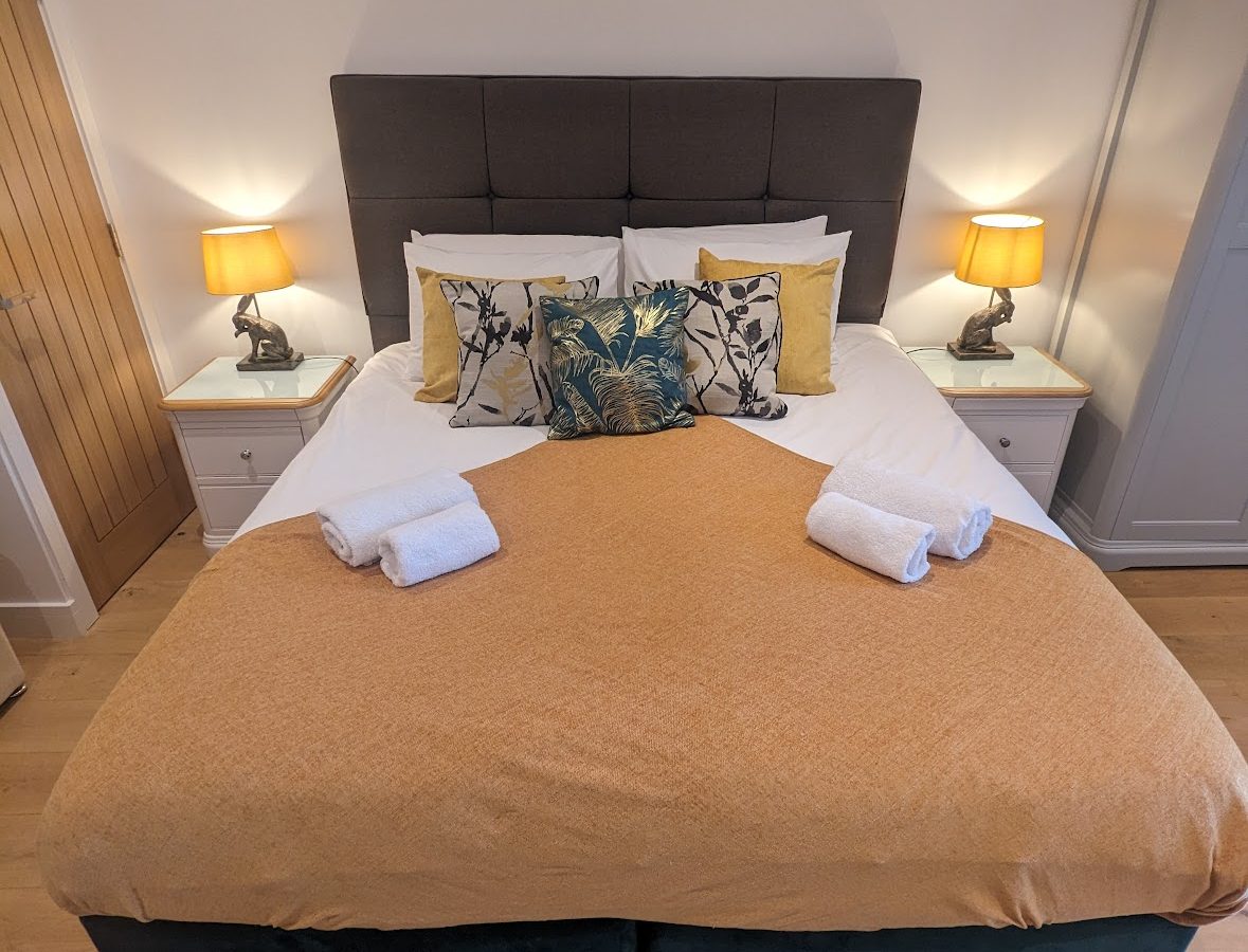 A neatly made bed with orange bedding, white pillows, and two neatly rolled towels enhances the warm ambiance of Oakwood House. Two lamps on bedside tables are lit.