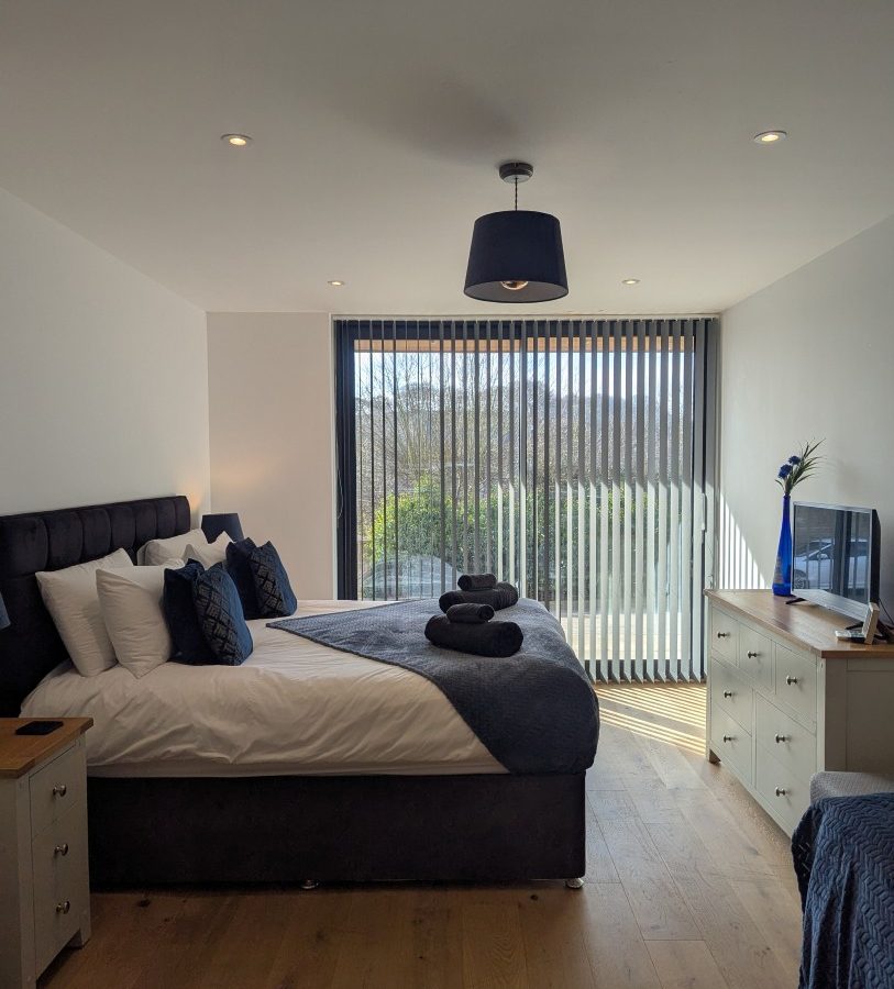 A modern Oakwood House bedroom with a large bed, blue linens, TV-topped chest of drawers, and vertical blinds.