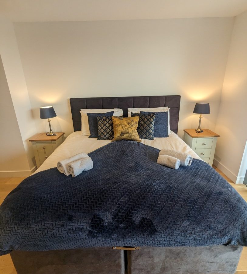 In the Oakwood House, a neatly arranged bed with blue bedding and decorative pillows is flanked by two nightstands with lamps, adding charm to the modern bedroom.