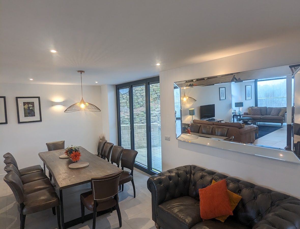 The modern dining and living area at Oakwood House features a table, chairs, sofa, mirror, and expansive windows that offer a view of the rustic stone wall.