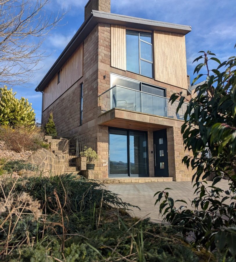 Oakwood House: A modern two-story home with large windows, balcony, brick exterior, surrounded by lush plants and clear blue sky.