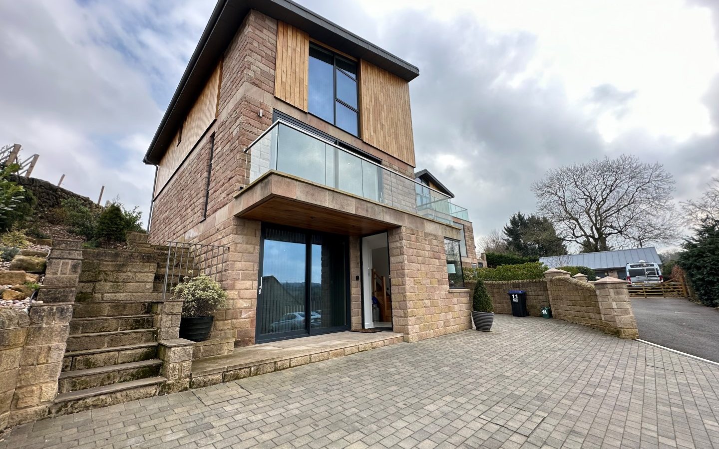 Oakwood House, a modern two-story residence, features large glass windows, a stone facade, and a paved driveway. Surrounded by trees under an overcast sky, it exudes contemporary elegance amidst natural beauty.