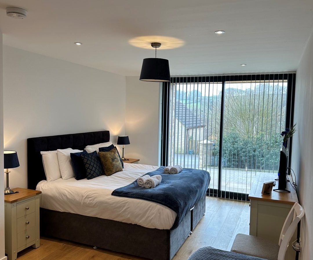 Modern bedroom in an Oakwood house featuring a double bed with blue bedding. Enjoy the view through large windows, complemented by bedside tables and lamps.