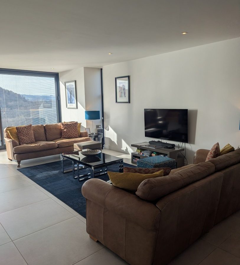 Modern living room in Oakwood House, featuring two brown sofas, a wall-mounted TV, and a coffee table on a dark blue rug.