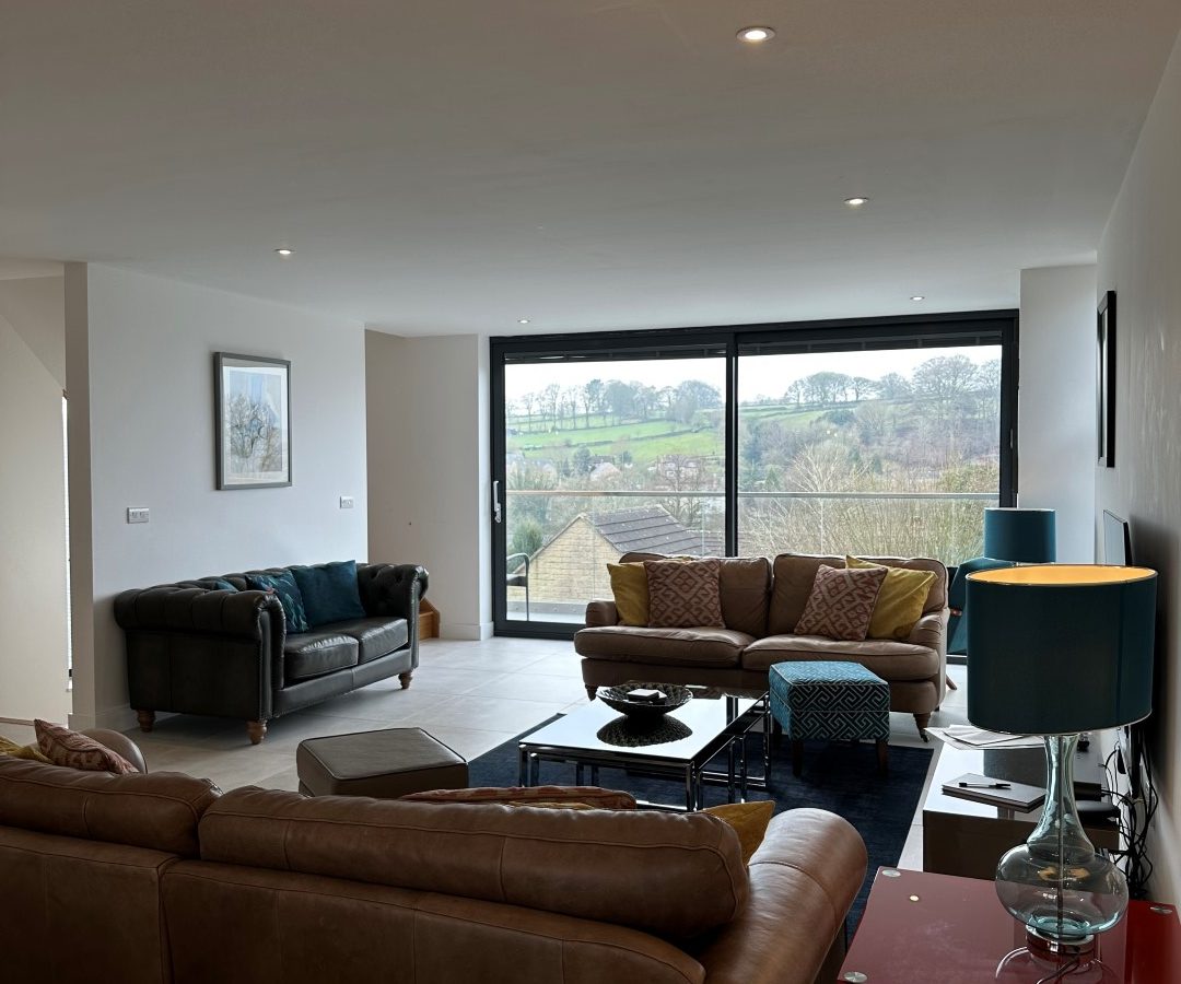 The modern living room at Oakwood House features stylish sofas, a sleek coffee table, and elegant lamps, all illuminated by natural light streaming through large windows that offer a breathtaking view of the lush green landscape outside.