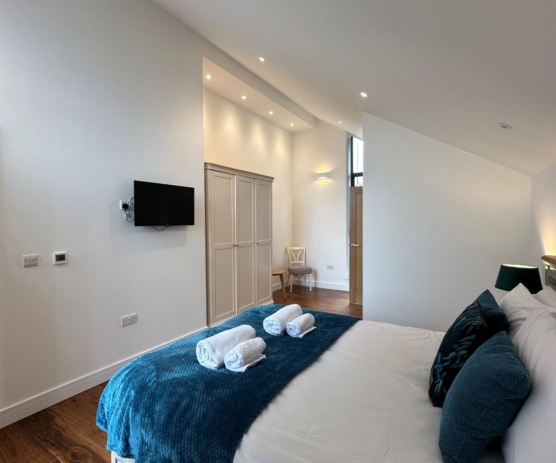 The modern bedroom at Oakwood House features pristine white walls, a bed adorned with blue accents, a wall-mounted TV, a spacious wardrobe, and elegant wooden flooring.