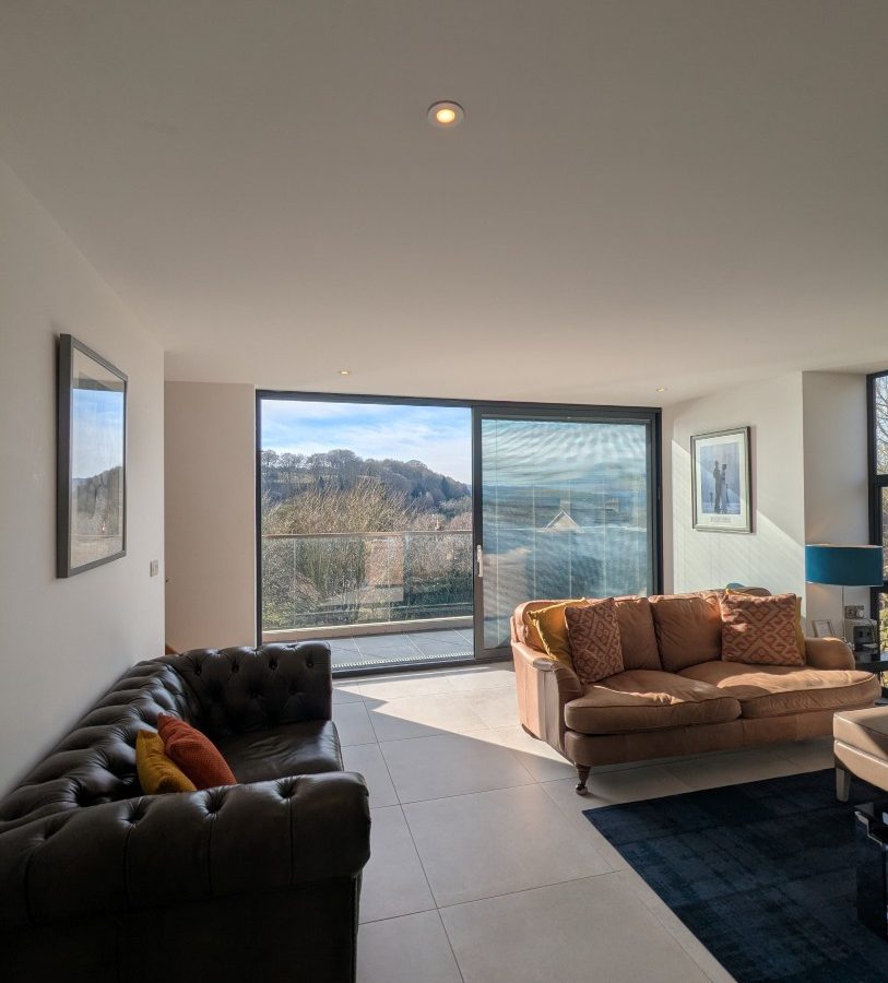 Bright living room at Oakwood House with large windows, a dark sofa and light couch, overlooking a hilly landscape.