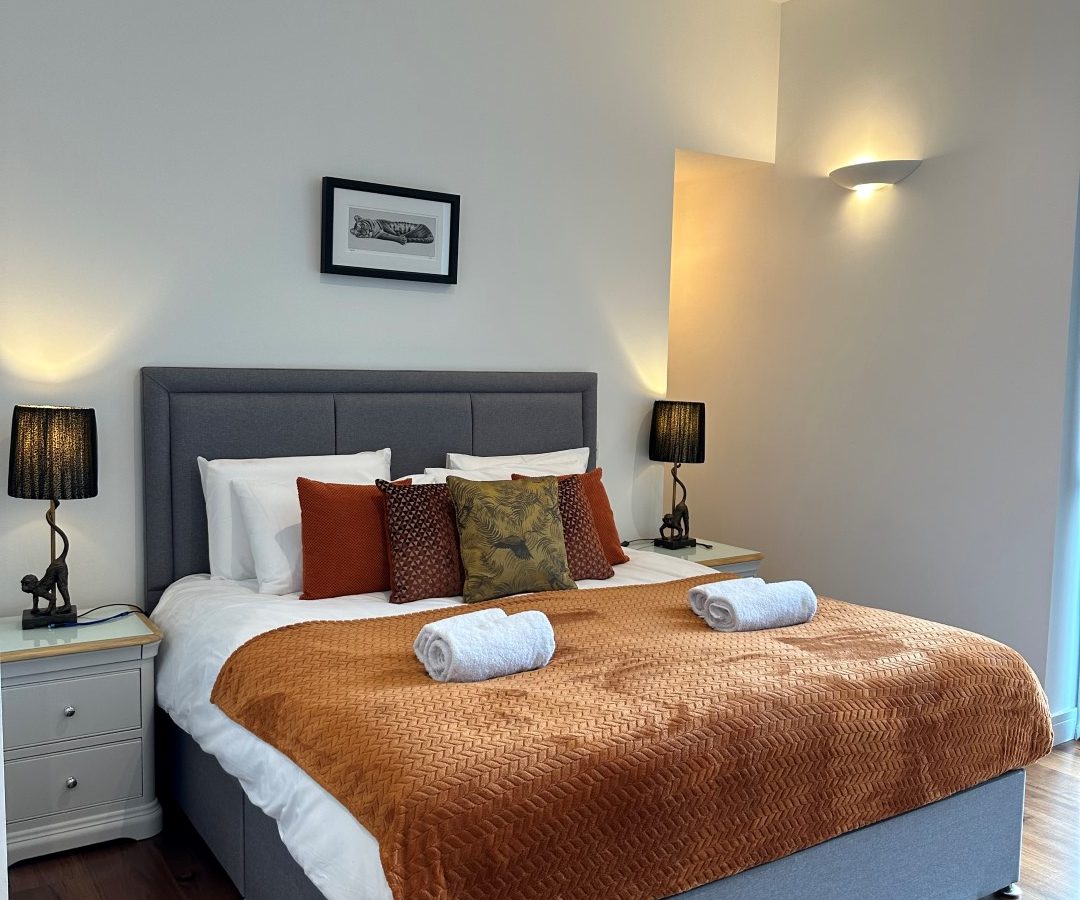 The Oakwood House features a modern bedroom with a gray bed and vibrant orange bedding, flanked by two sleek bedside lamps. The pristine white walls complement the warm wooden floor, creating an inviting and stylish retreat.