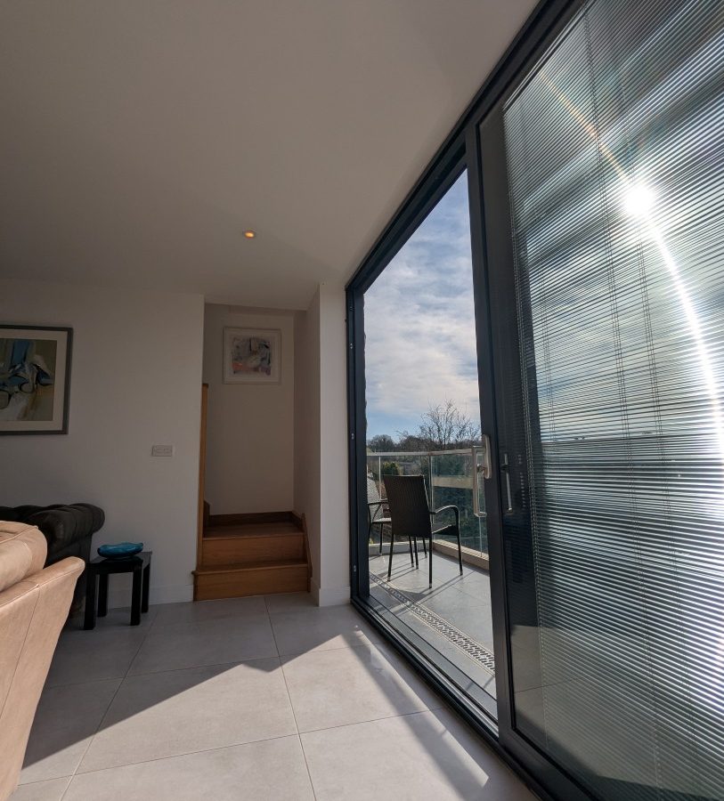 Bright room at Oakwood House with large glass doors leading to a balcony, a sofa on the left, and a small staircase in the corner.