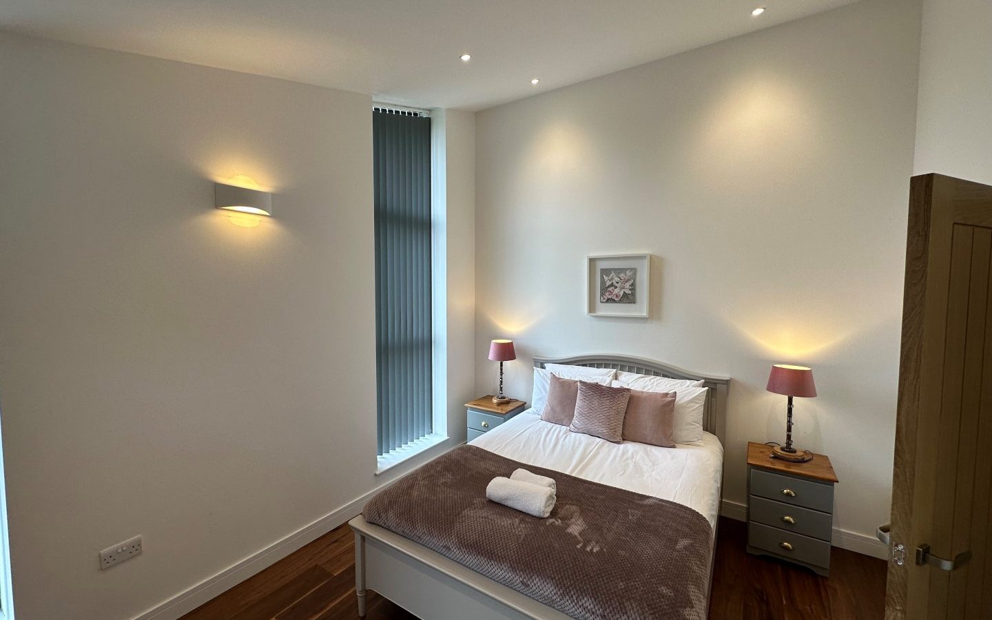 The minimalist bedroom at Oakwood House features a neatly made bed adorned with brown pillows and a blanket, flanked by two bedside tables with lamps and complemented by elegant wall lights.