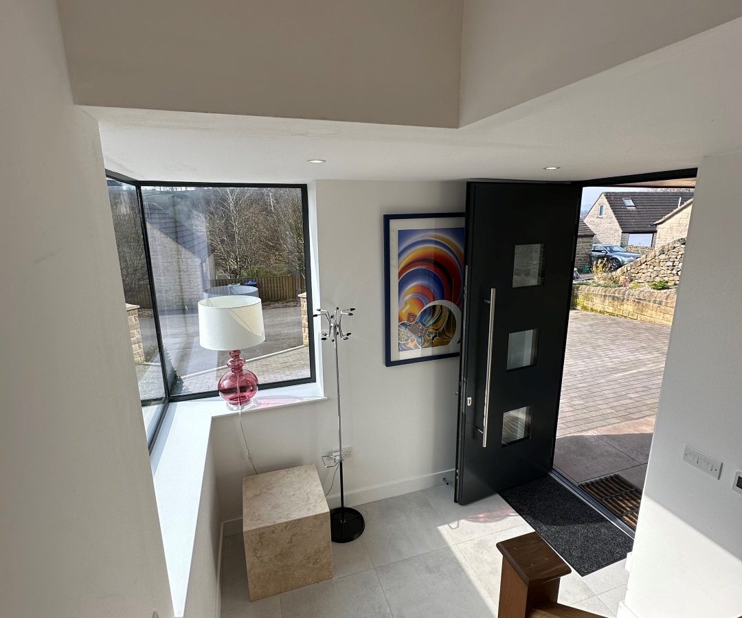 The modern entryway of Oakwood House features a sleek glass door, an abstract painting, and a lamp resting on a cube table near the large window, all complemented by the elegant wooden staircase.