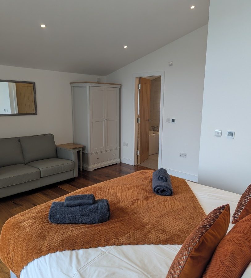 Cozy bedroom in Oakwood House, featuring a bed, brown and gray accents, a gray sofa, wardrobe, and an open bathroom door.