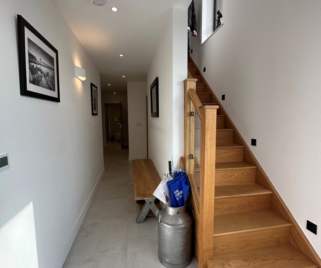 The modern hallway of Oakwood House features a wooden staircase, wall-mounted pictures, a bench, a grey floor, and a silver milk churn cleverly used for storage.