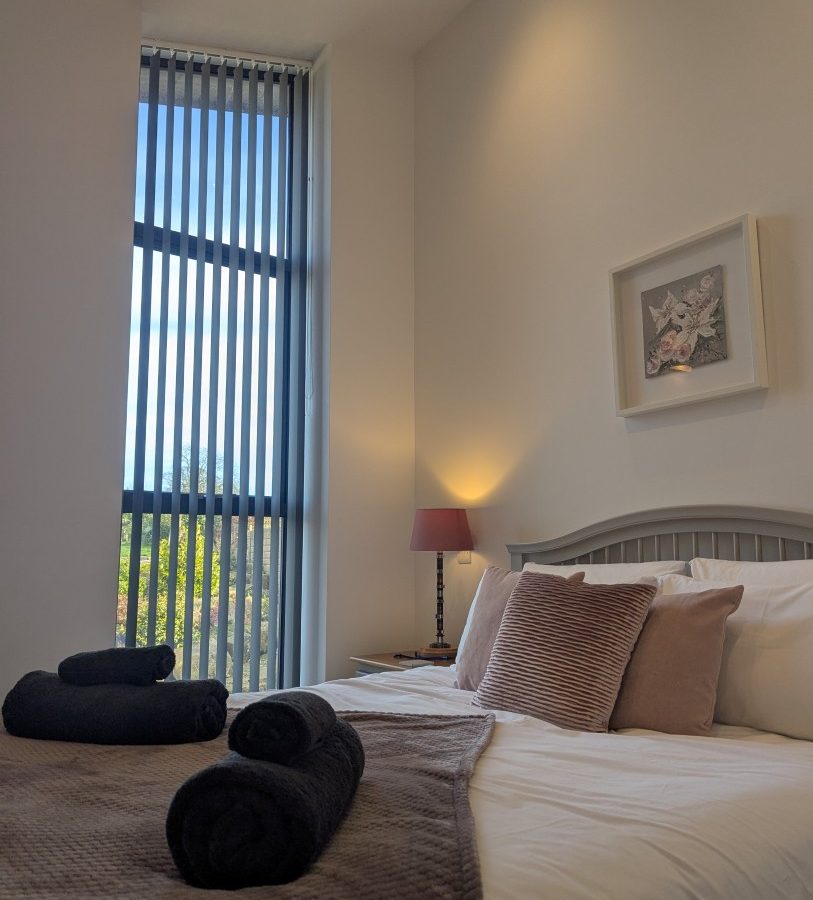 A bedroom at Oakwood House features a neatly made bed, rolled towels, a side table with a lamp, and tall vertical blinds.