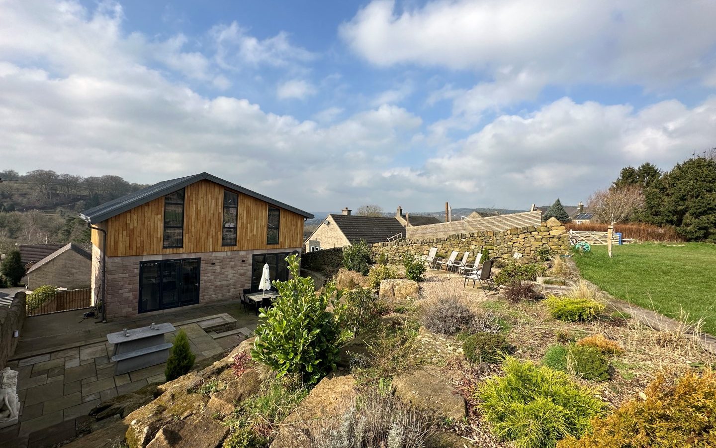 The Oakwood House, a modern residence with wooden accents, is nestled amid a flourishing garden enclosed by a stone wall under a partly cloudy sky.