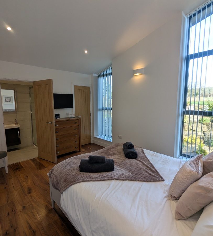 Modern bedroom at Oakwood House with a double bed, pillows, tall windows, wooden floor, and open door to the bathroom.