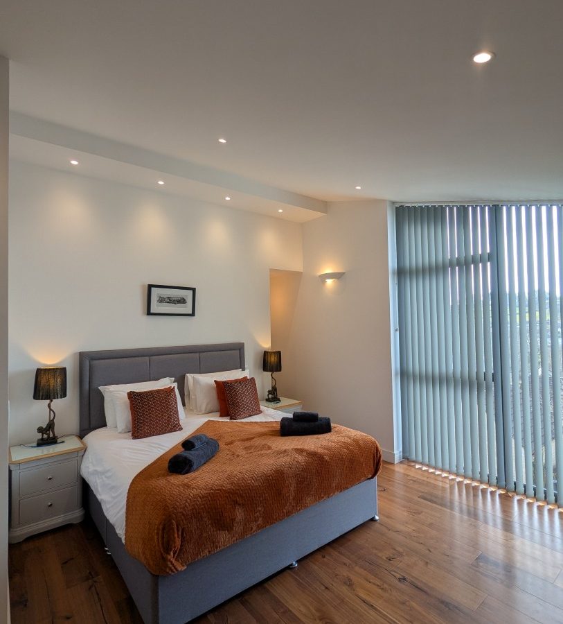 Modern bedroom at Oakwood House with a double bed, orange throw, dark lamps on side tables, wood floor, and tall vertical blinds.