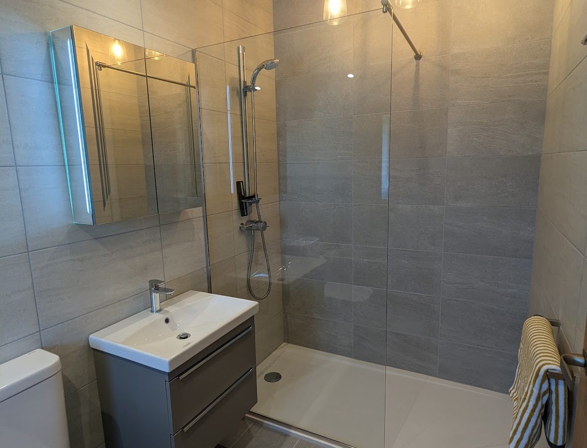 The Oakwood House features a modern bathroom with a sleek glass shower enclosure, wall-mounted sink, and toilet, all illuminated by three stylish pendant lights overhead.