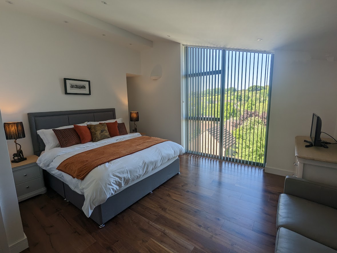 The Oakwood House features a bright bedroom with a large bed, orange accents, wooden floor, and a window with blinds offering a scenic view.