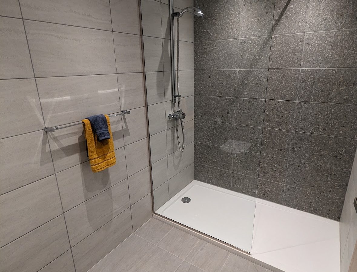 Modern bathroom at Oakwood House with tiled walls and floor, featuring a glass shower enclosure, towel on rail, and a toilet in the corner.