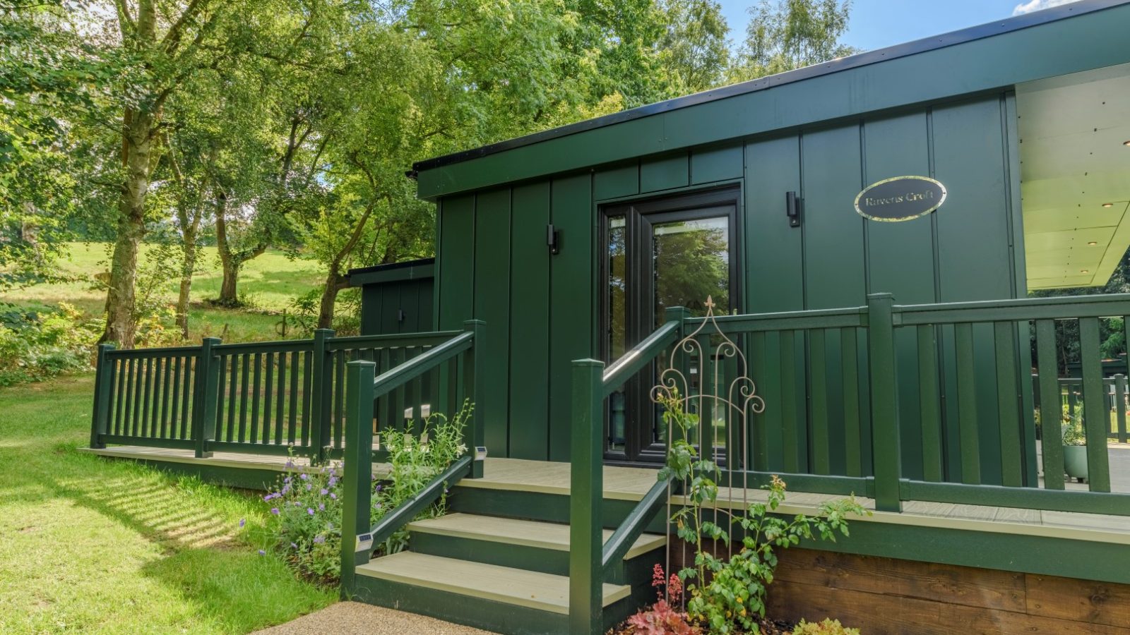 A small, dark green cabin at Rowley Meadow, with steps leading to a door, surrounded by trees under a clear blue sky.