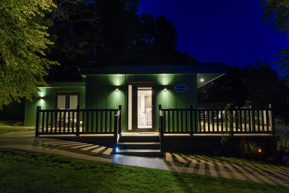 A charming green house in Rowley Meadow, with a well-lit porch at night, surrounded by trees and featuring a wooden deck with steps.