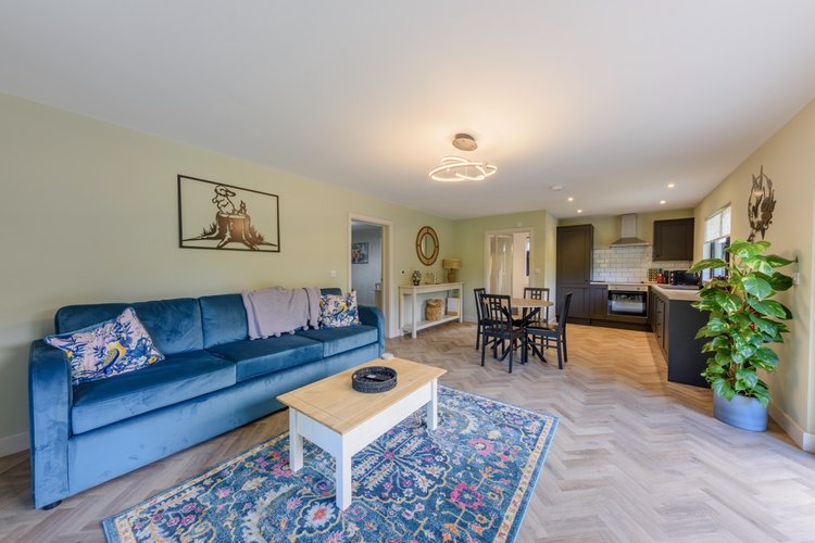 The living room at Rowley Meadow features a blue sofa, wooden coffee table, and a patterned rug, seamlessly flowing into the dining area with an open kitchen in the background.