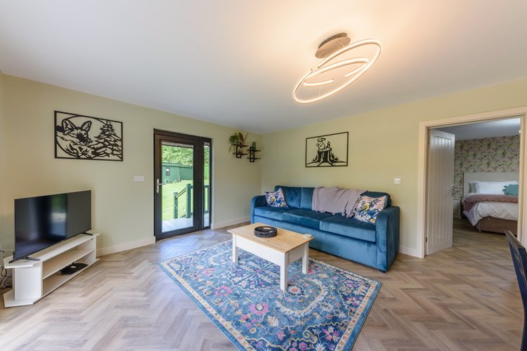 A modern living room in Rowley Meadow features a blue sofa, TV, wooden coffee table, and decorative rug, leading to an open door and a bedroom.