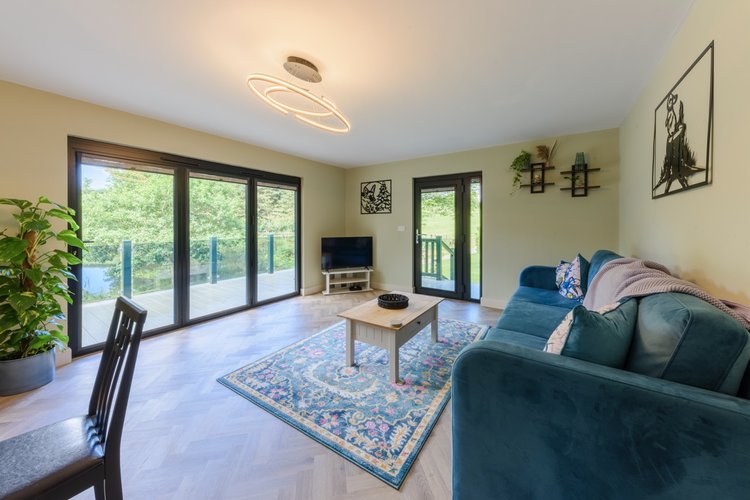 The modern living room features a blue couch, coffee table, and TV, with large windows offering a view of the lush greenery beyond Rowley Meadow.