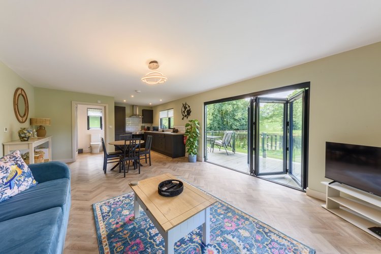 The spacious open-plan living area at Rowley Meadow features a comfortable sofa, stylish coffee table, elegant dining set, and modern kitchen, all complemented by large glass doors that lead outside.