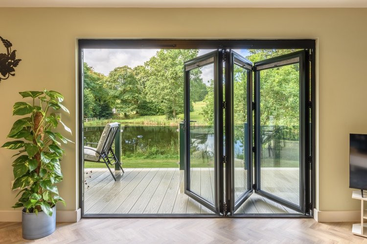 Glass doors open to a deck with chairs, offering a picturesque view of Rowley Meadow's pond, surrounded by trees. A potted plant is near the door.