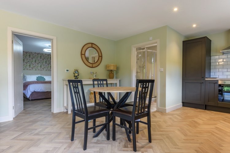 The Dining area in Rowley Meadow features a round table with four chairs, seamlessly connected to the kitchen. A door leads to the bedroom, all enveloped in calming neutral tones.