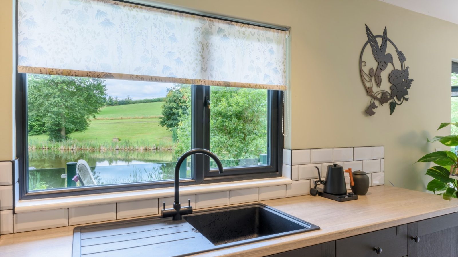 Kitchen sink with matte black fixtures under a large window revealing Rowley Meadow. Electric kettle on the wooden countertop.