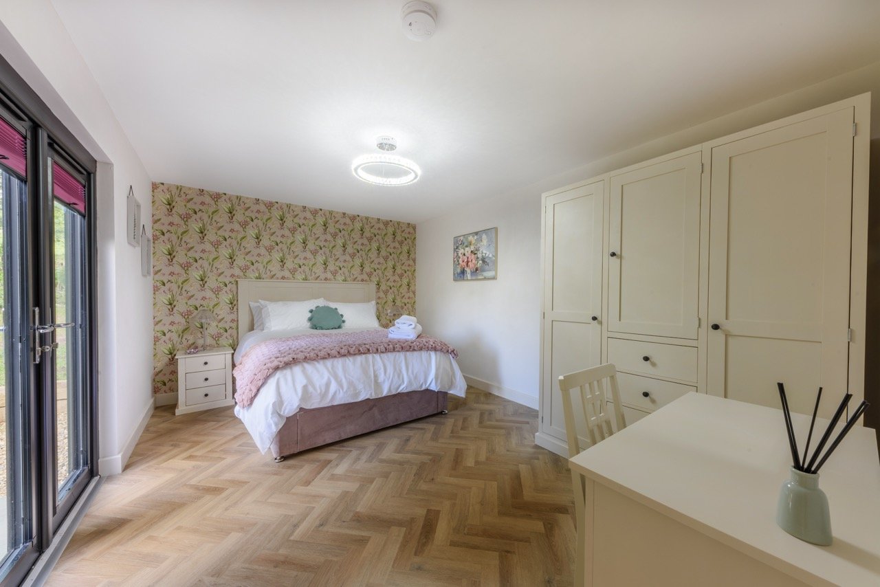 The bedroom in Rowley Meadow boasts a charming floral accent wall, complemented by a cozy bed draped with a pink blanket. A sleek white wardrobe and desk add functionality, while a large window lets natural light flood the serene space.