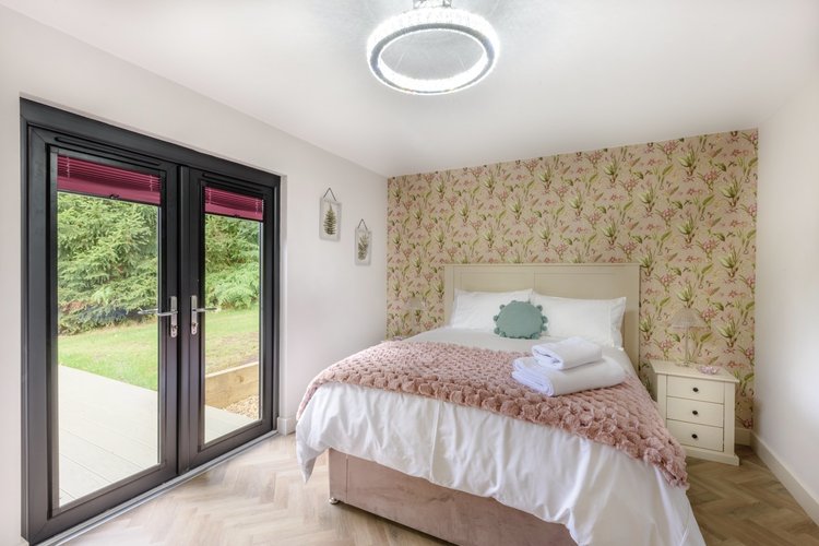 A cozy bedroom with a floral accent wall and a neatly made bed opens up through glass doors to a stunning view of Rowley Meadow.