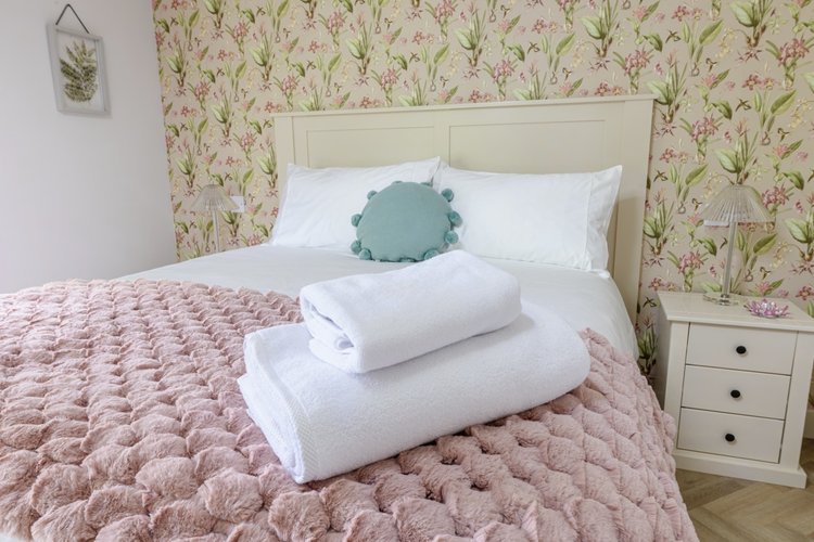 The bedroom features a charming floral wallpaper reminiscent of Rowley Meadow, with a bed adorned in crisp white linens topped by a pink textured blanket. Two neatly folded towels complete this serene setting.